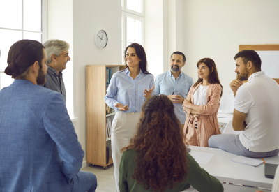 Group of happy corporate employees having an interesting meeting with a female team leader or business coach. Smiling young woman talking to a group of people during a workshop in a modern office