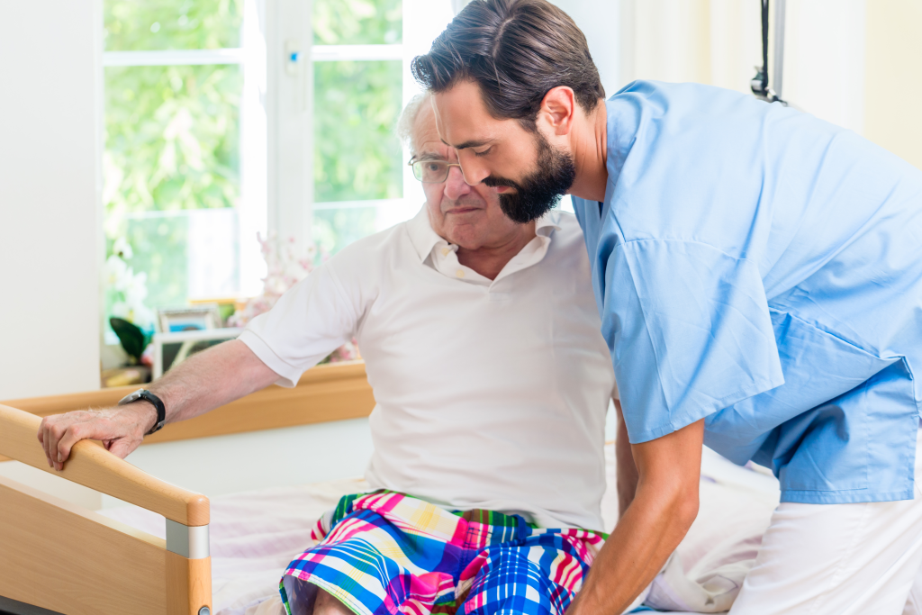 Altenpfleger hilft älterem Mann aus Rollstuhl ins Bett